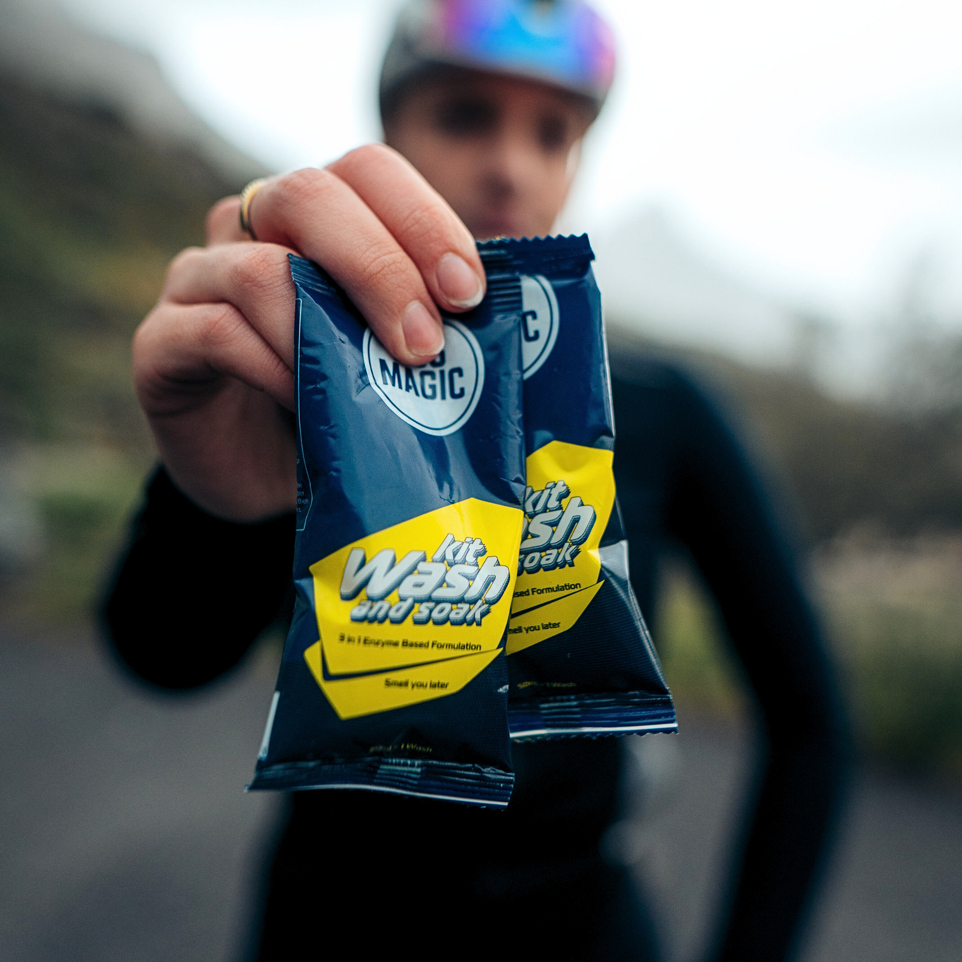 Cyclist holding up two sachets of ASS MAGIC Kit Wash and Soak, a portable sports kit wash designed for odour control and easy gear cleaning.