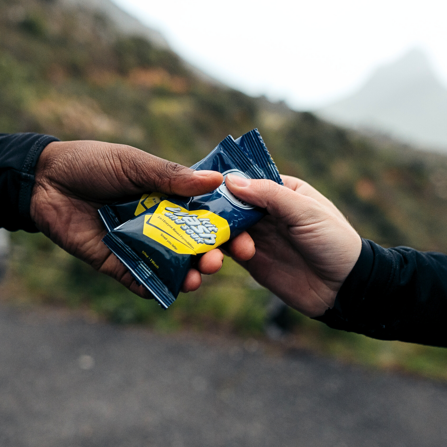 Close-up of hands exchanging sachets of ASS MAGIC Kit Wash and Soak, a convenient travel-sized solution for washing sports kit and controlling odour on the go.